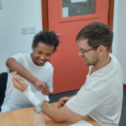 One man practises bandaging another man's arm, they're both sitting a desk smiling as they do this